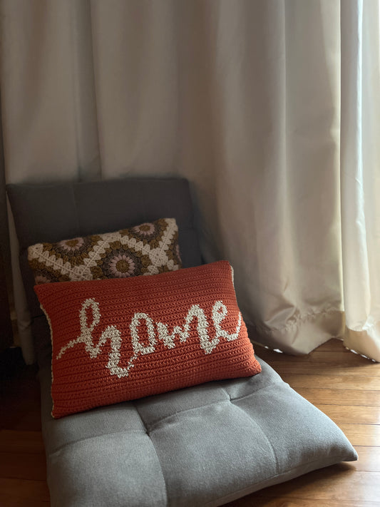 Handmade crochet pillow in burnt orange with 'Home' text, placed on a gray cushion in a room setting