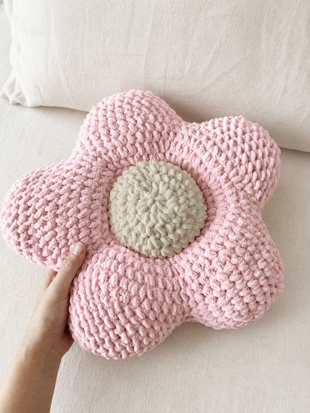 A hand holding a pink crocheted flower-shaped cushion with a beige center, set against a soft cream fabric background.